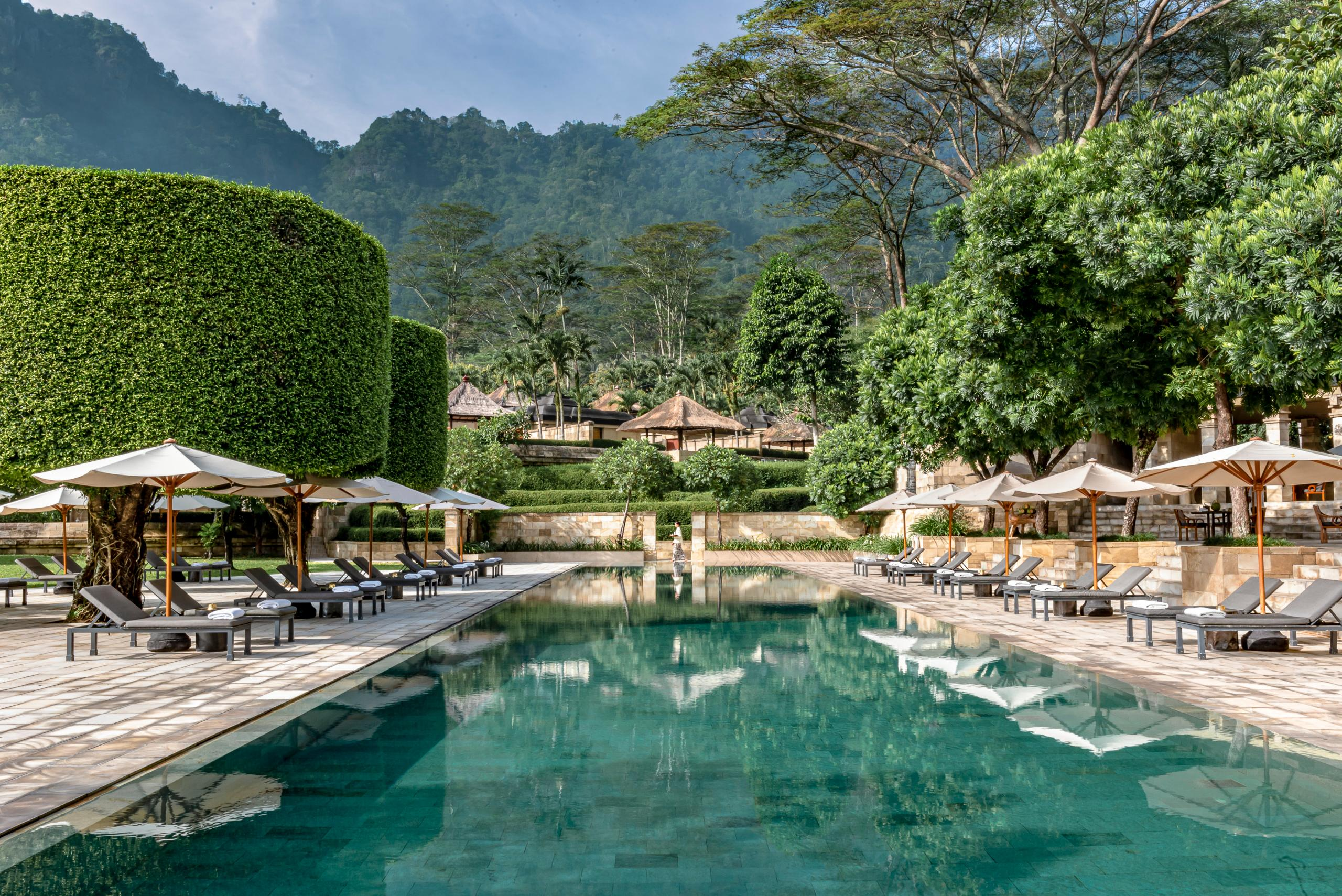 Resort swimming pool lined with loungers and white umbrellas, surrounded by tropical gardens and forested hills.