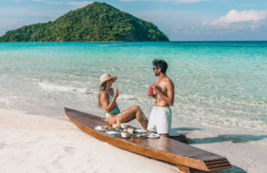 Couple enjoying a beach picnic at Bawah Reserve, Indonesia, beside turquoise water.