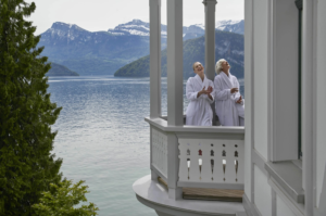 Two guests in bathrobes laughing on a balcony at Chenot Palace Weggis, overlooking Lake Lucerne and the Swiss mountains.