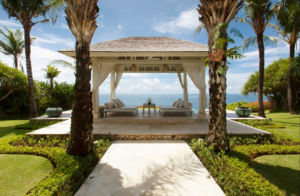 Ocean-view pavilion at The Ungasan, Bali, with white lounge seating framed by palm trees and tropical gardens.